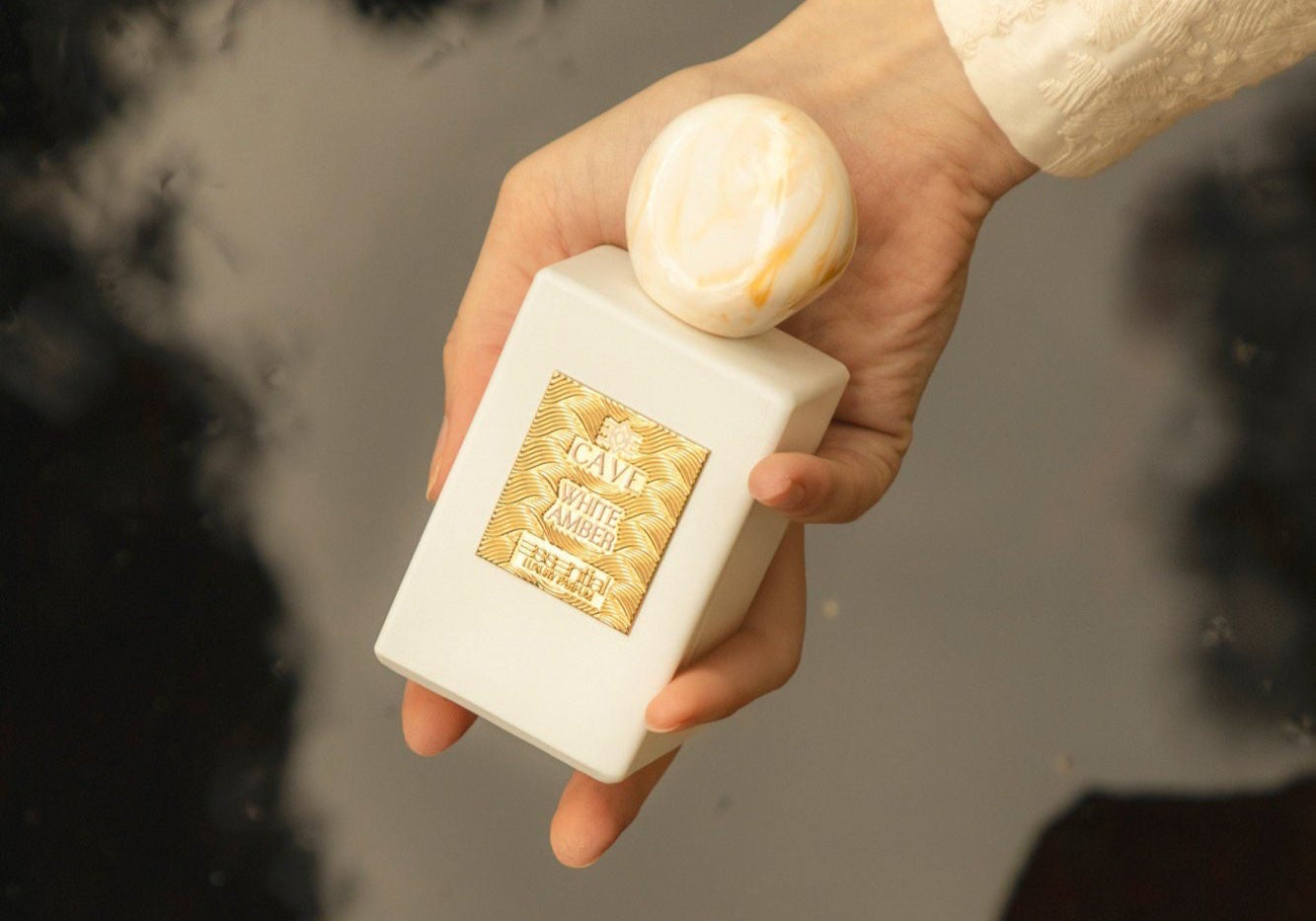 Hand holding a white bottle with a gold label over pink flowers on a reflective surface