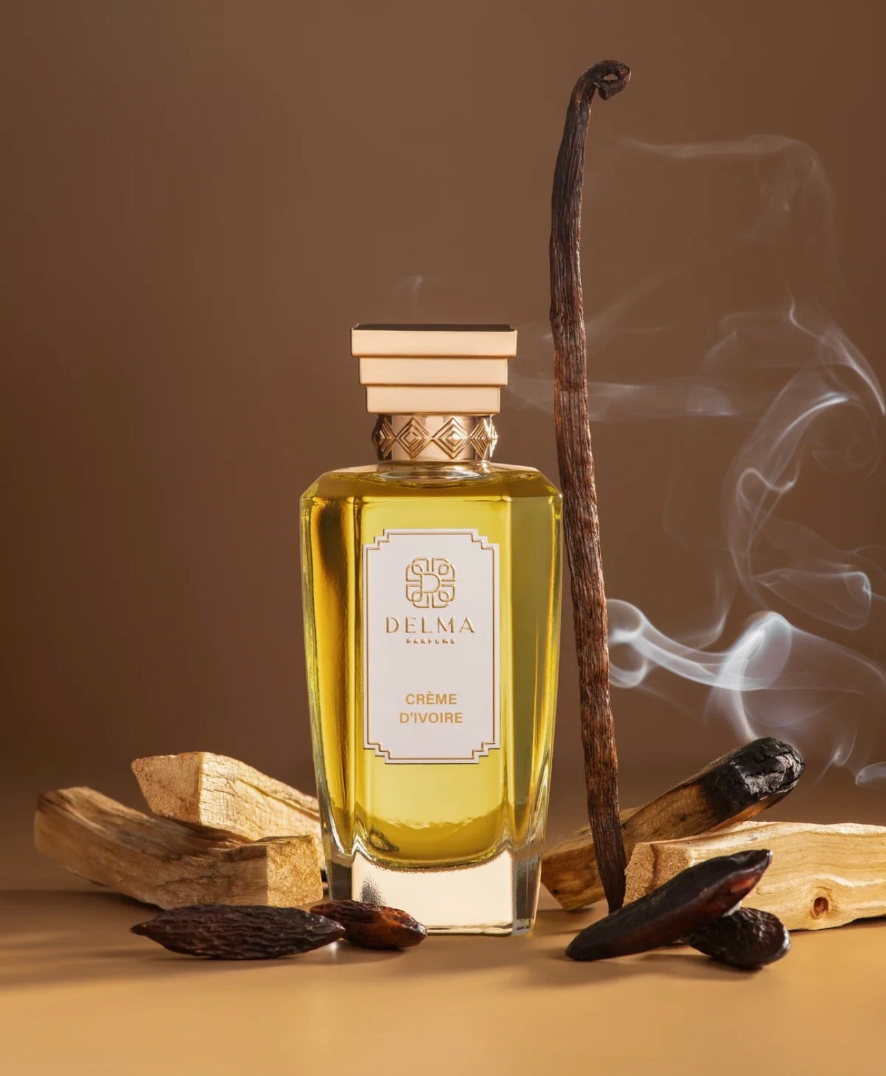 A bottle of DELMA Paris Crème Dôgne perfume with golden-yellow liquid, set against a warm brown background with vanilla pods, wood pieces, and a smoking vanilla pod.