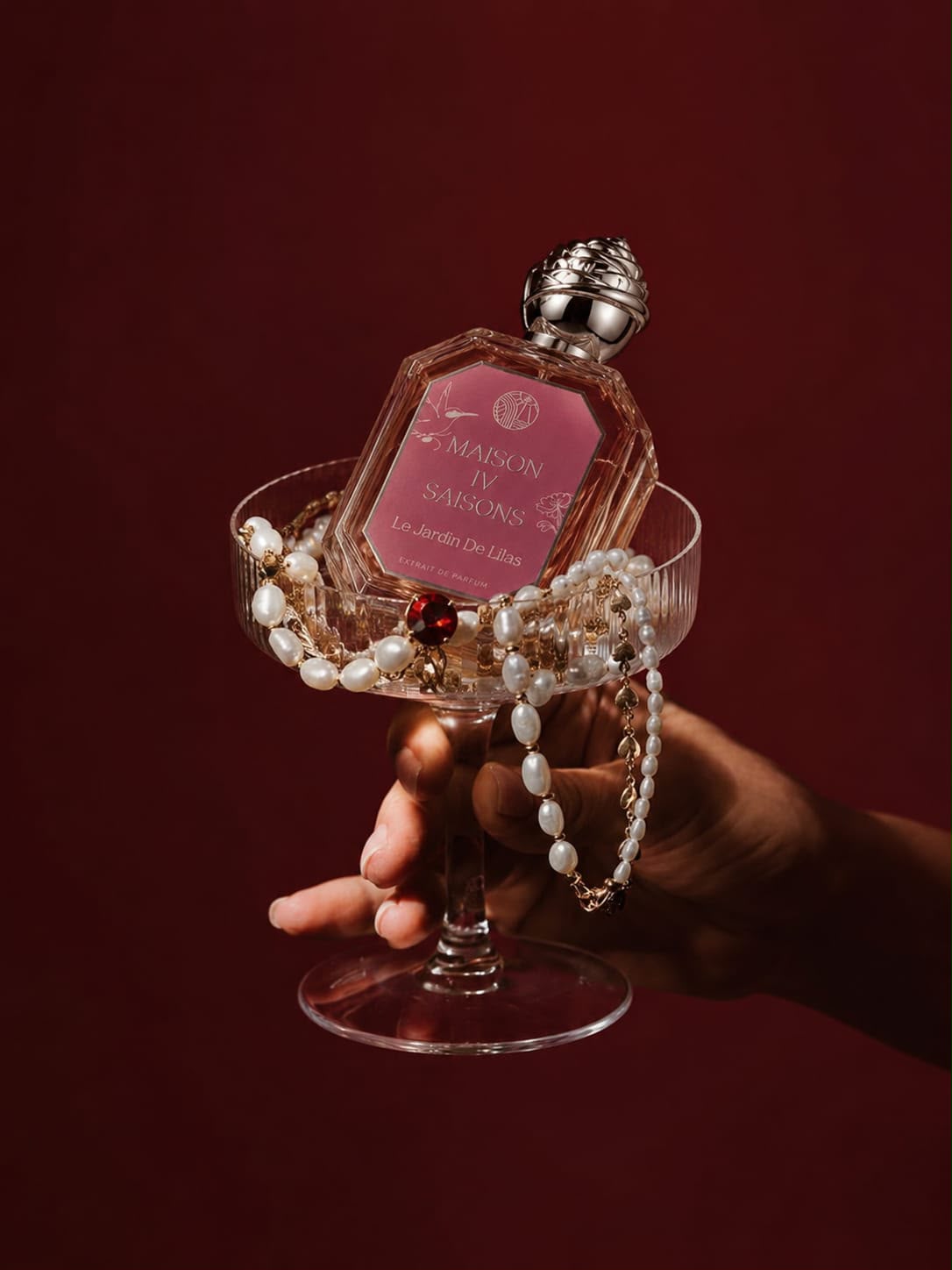 A hand holds a clear stemmed glass containing a pink perfume bottle labeled Maison de Marsons, wrapped with white pearl strands and small red jewel accents, against a dark burgundy background.