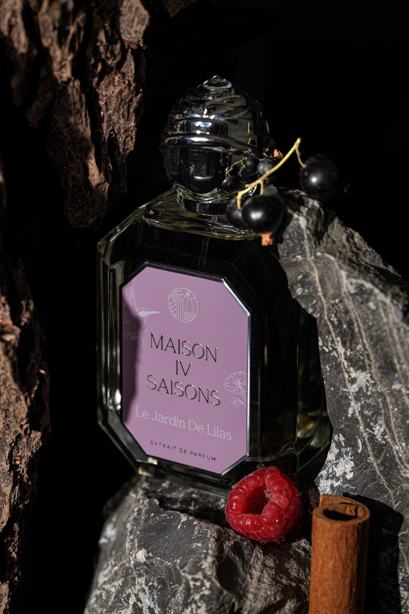 A clear glass perfume bottle with a silver cap and a lavender label reading 'Maison IV Saison Le Jardin De Lilas', styled on dark rocks with a raspberry, cinnamon stick, and small black berries against a dark background.