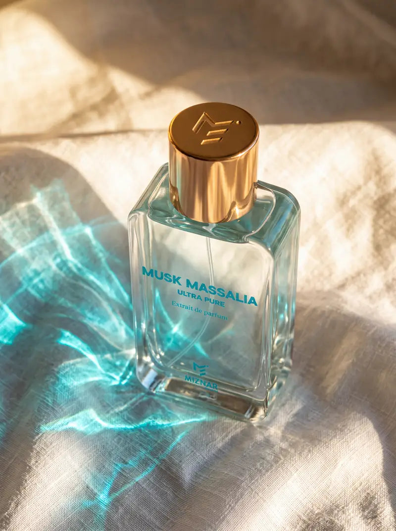 A clear glass perfume bottle labeled 'Musk Massalia Ultra Pure' with a metallic gold cap, resting on a textured beige fabric under warm sunlight and blue shadow play.