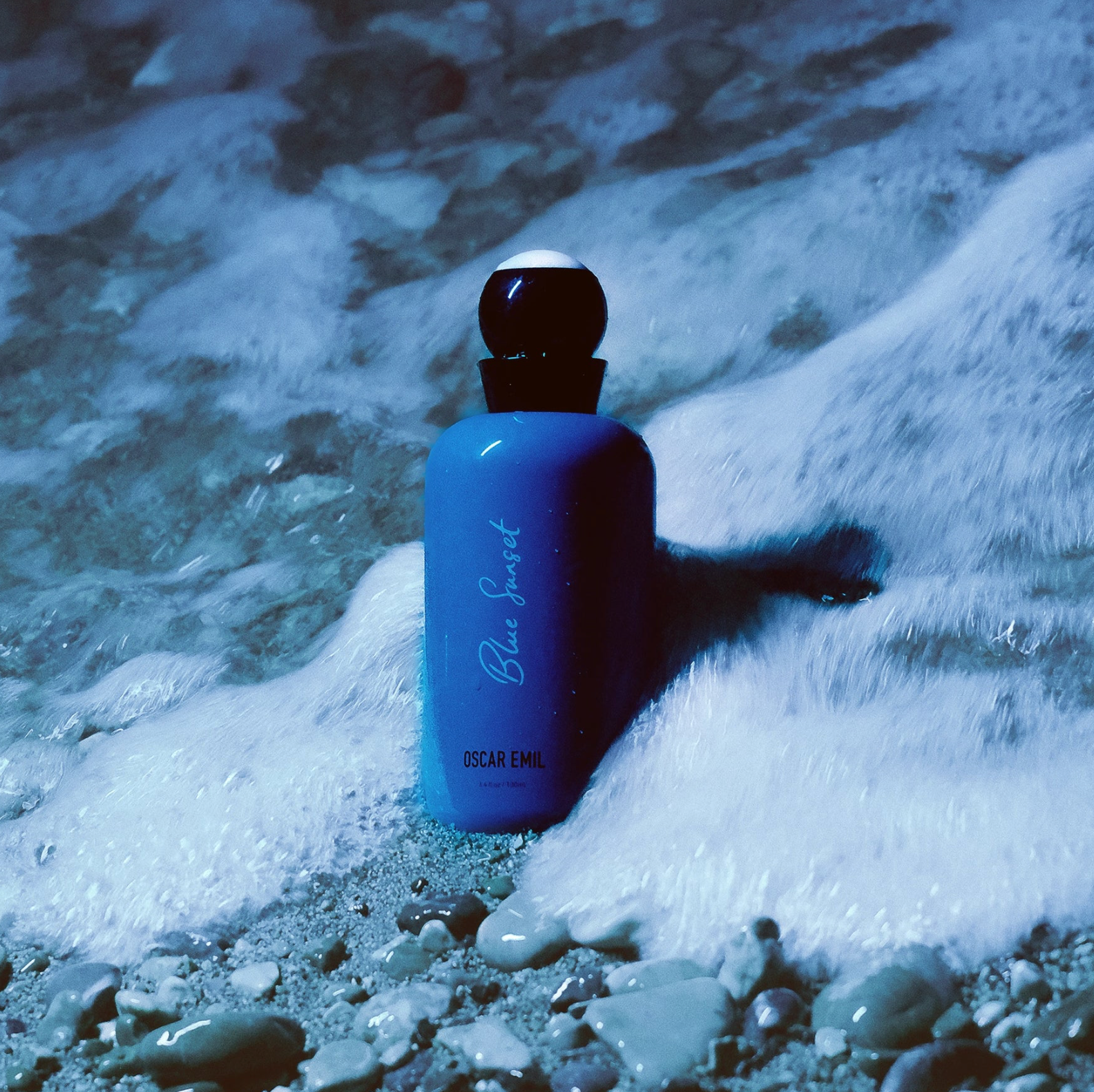Blue perfume bottle labeled 'Blue Sunset' by Oscar Emil on a rocky surface with water.