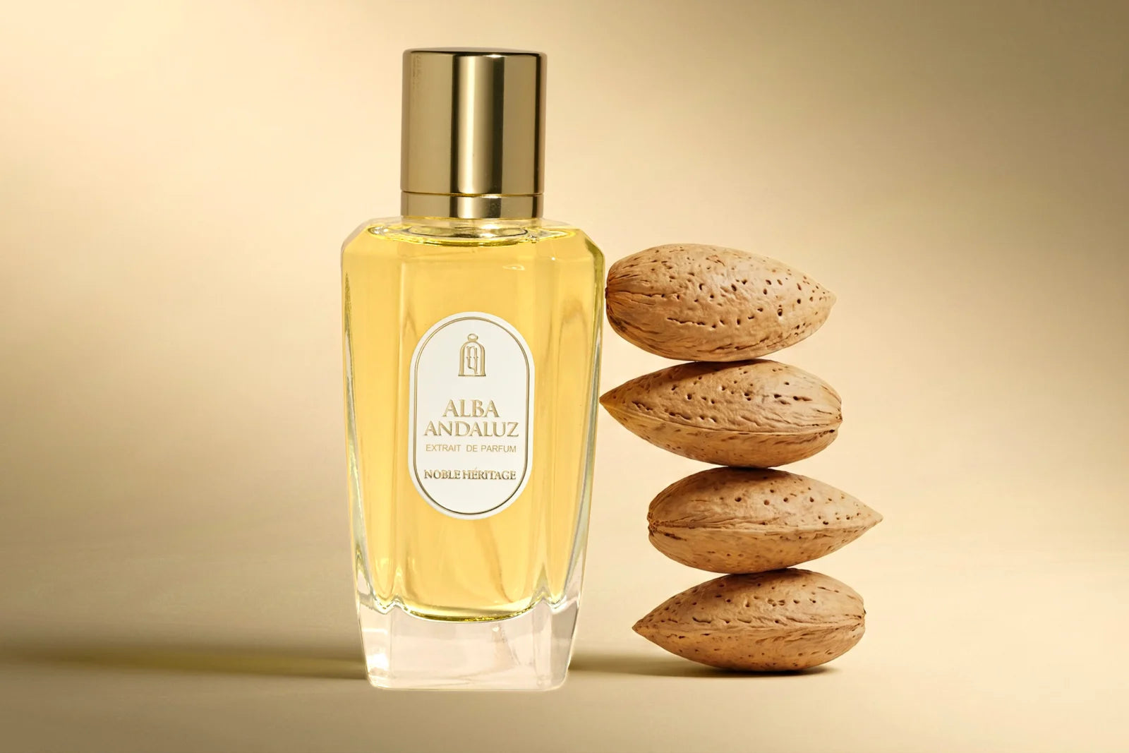 A clear glass perfume bottle filled with pale golden-yellow liquid, topped with a metallic gold cap, standing beside a stack of four whole almonds on a warm beige background. The bottle label reads Alba Andaluz.