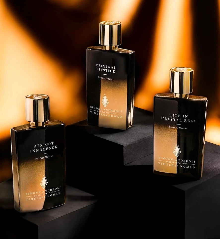 Three dark glass perfume bottles with gold caps on black stands against a warm, blurred background.